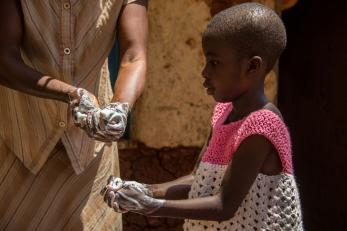 Young girl wearing pink and white crocheted top washing her hands with soap. another person in background is washing their hands too.