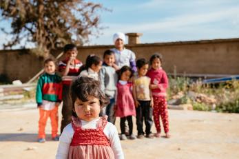 A group of children in Lebanon smiles and snickers in the background as one young girl approaches the camera inquisitively 