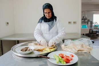 A woman wearing glasses and a headscarf prepares lunches with meat and vegetables in a commercial kitchen