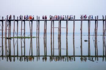 Pedestrians in colorful clothing walk across a wooden bridge elevated on pillars and reflected in the water beneath it