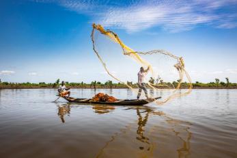 Fishermen in a small wooden boat cast a large yellow net against a blue sky