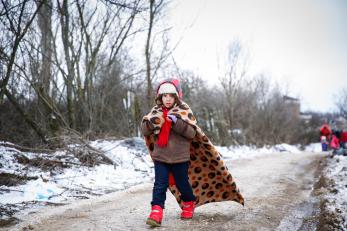 A girl in a jacket, boots, scarf and hat walks down a snowy road with a blanket wrapped around her shoulders.