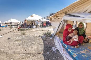 Two women sit beneath a white canvas tent. One is holding and feeding a baby on her lap.