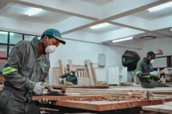 A person uses wood working tools while wearing a face mask in a room with other face masked workers.