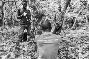 A person stands in a wooded area using a mobile device while another person sits on the ground and a third person stands nearby smiling.