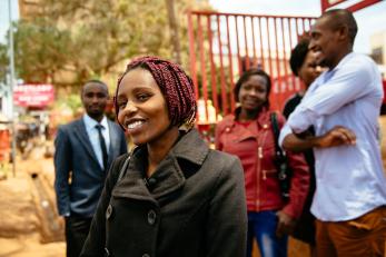 A smiling person in a black coat wears a lapel microphone while a group of smiling people stand behind her in Kenya.
