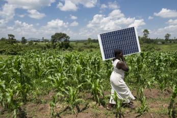 A person walks along a field with a solar panel on their shoulder.