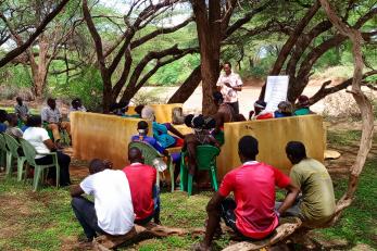 Kenyan community members meet in the outdoors under trees.