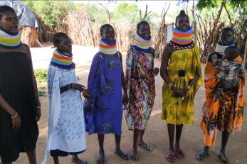 Six Kenyan women, one of whom holding a toddler, stand together in their village.