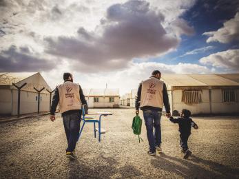 Two Mercy Corps team members and a small boy walking; one man is carrying a chair and the other man is carrying the boy's backpack and walking hand-in-hand with the boy.