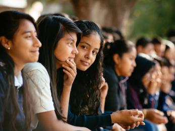 School children sitting in a row, one smiles at the camera.