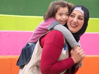 An adult smiles while carrying a smiling child clinging to their back.