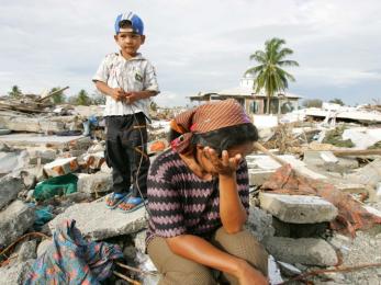 Small boy stands on rubble in background. in foreground, a seated woman wipes away tears.