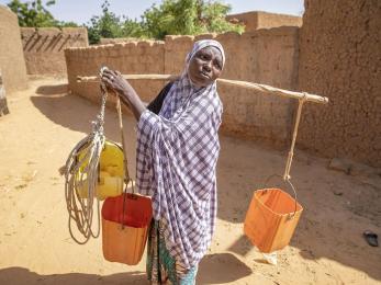 Halma, pictured in a purple and white shawl carrying three buckets balanced across her shoulders on a large stick