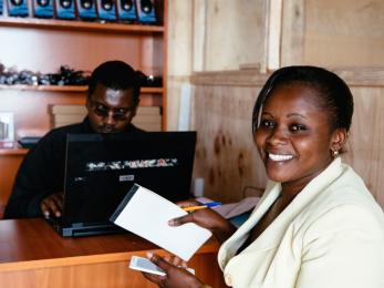 Lucy at a counter across from a man on a computer, smiling and holding a cell phone, notepad and pen.