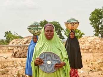 Three nigerian woman in traditional dress; two with balanced loads on their heads.