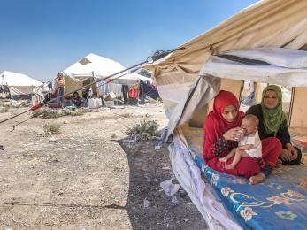 Two women sit beneath a white canvas tent. one is holding and feeding a baby on her lap.