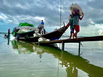 People carry fishing gear from a boat docked under a cloudy sky.