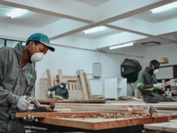 A person uses wood working tools while wearing a face mask in a room with other face masked workers.