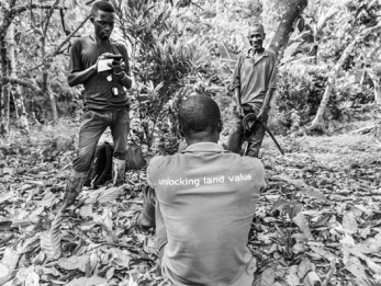 A person stands in a wooded area using a mobile device while another person sits on the ground and a third person stands nearby smiling.