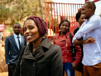 A smiling person in a black coat wears a lapel microphone while a group of smiling people stand behind her in kenya.