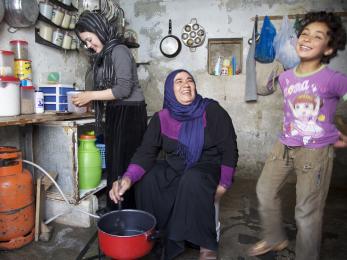 An adult stirs a pot while smiling at a happy child while another adult preps ingredients nearby.