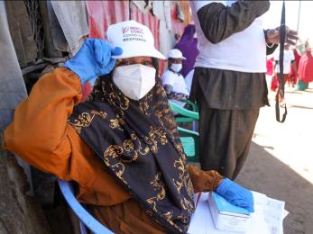 A person wearing face mask shows off their mercy corps hat to the camera on a busy street in somalia.