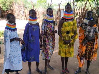 Six kenyan women, one of whom holding a toddler, stand together in their village.