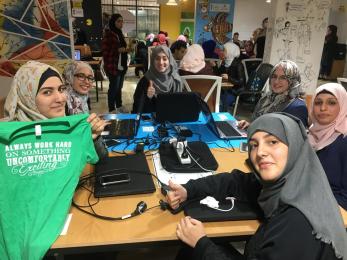 A group of young people sit a table with laptops and mobile devices and pose for the camera.