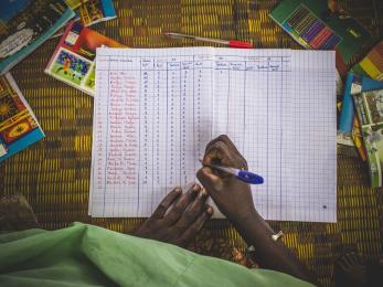 A hand holding a pen marks a paper spreadsheet.