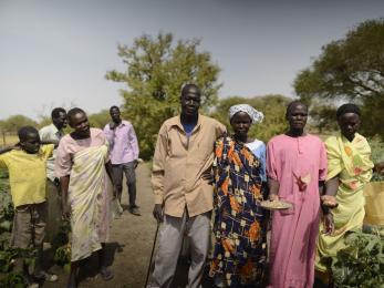 A group of people stand  outside together holding harvested agricultural goods in south sudan. 