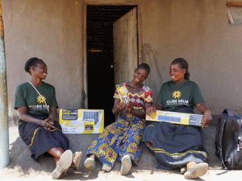 Ugandan women on front porch of home with new, boxed solar panel systems.
